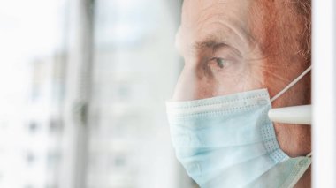 Maskeli, üzgün bakışlı emekli doğrudan pencereden bakıyor.