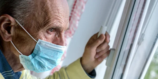 Homem idoso em azul máscara estéril admira paisagem urbana em casa — Fotografia de Stock
