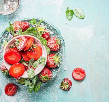 Dilimlenmiş çilekli kaseler pudra şekeri ve açık mavi kırsal arka planda nane yaprakları, üst manzara.