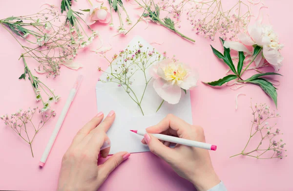 女性的手用铅笔写在开着的信封上 上面有花朵的排列 粉红桌子背景上的花店装饰设备 顶视图 问候和假日 — 图库照片