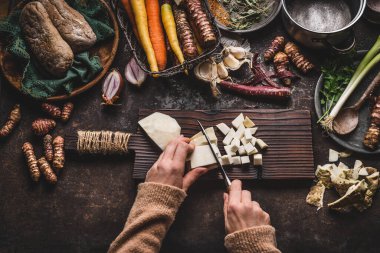 Koyu kırsal mutfak masasında çeşitli sebze ve çatal bıçaklarla kereviz doğrayan kadın elleri. Lezzetli sonbahar yemekleri için kök sebzeleri hazırlama, üst manzara. Sağlıklı yeme kavramı
