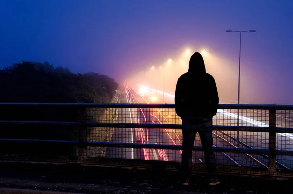 A hooded figure silhouetted against street lights. Looking down on a motorway with long expsoure traffic trails.