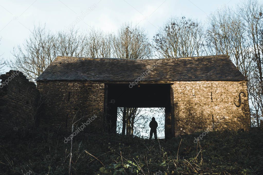 A sinister, silhouetted, hooded figure stands in a ruined old barn on a winters day. With a cold muted edit.