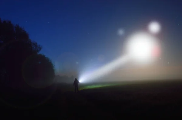 image of a silhouetted figure standing in a misty field at night watching a UFO in the sky, with a beam of light coming down.