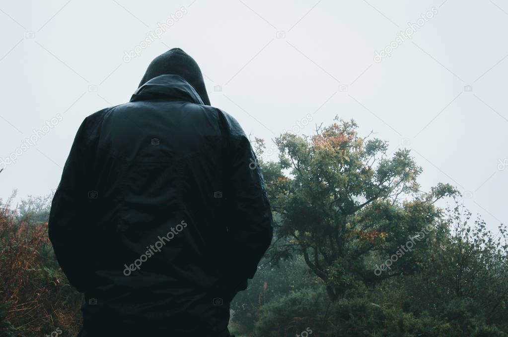 Looking up from behind at a mysterious hooded figure wet from rain on a country path.