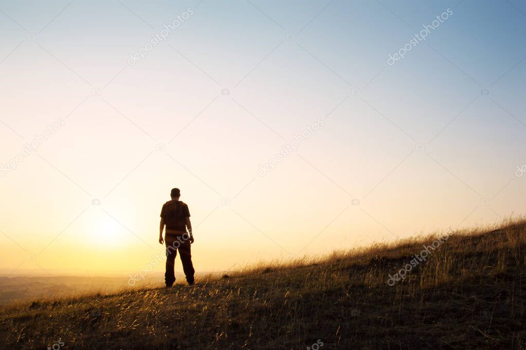 A silhouette of a lone hiker with sun flare, standing on a hill looking out towards the setting sun. 