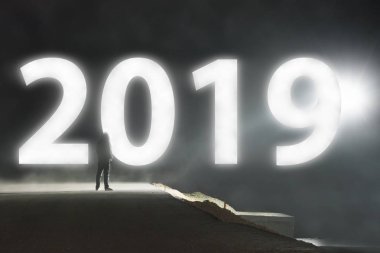 Yalnız bir kukuletalı rakam ürkütücü bir puslu yolda karanlıkta parlayan bir 2019 işaret bakarak geceleri ayakta.