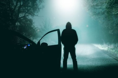 Yalnız, kapüşonlu şekil ayakta yanında bir puslu, ülke yolda araba farlar tarafından gece yukarıdan bir Ufo iniyor bir ışık demeti bakarak bir araba