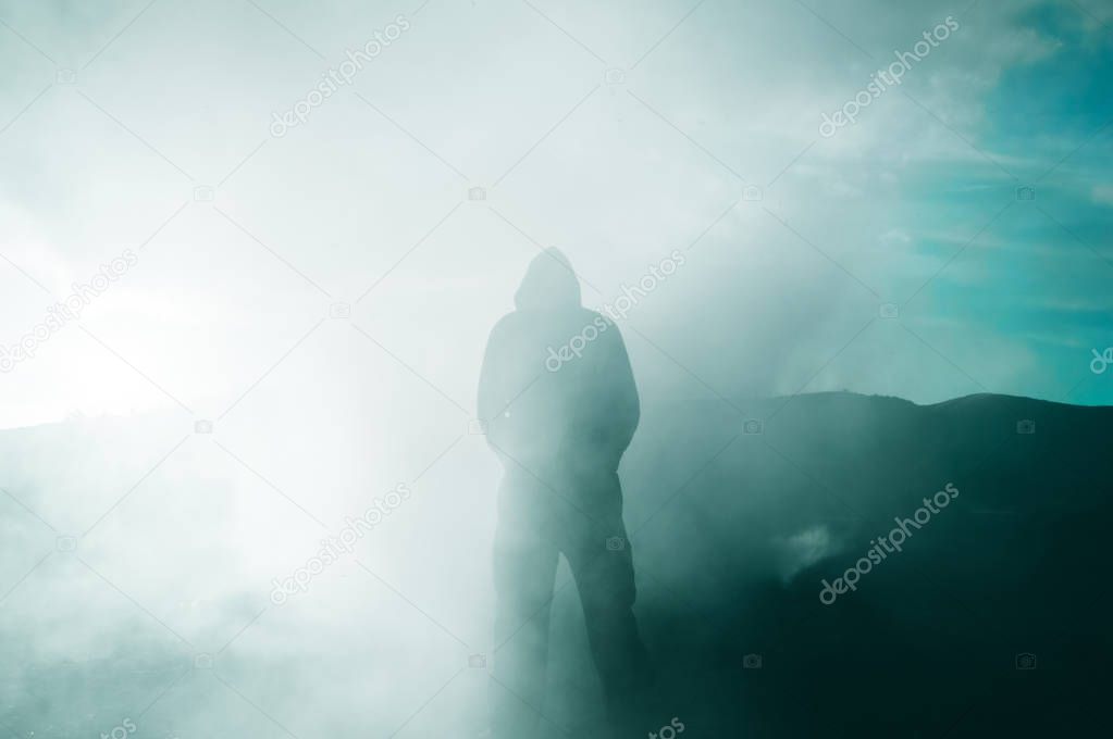 A hooded figure silhouetted by the sun as smoke surrounds him