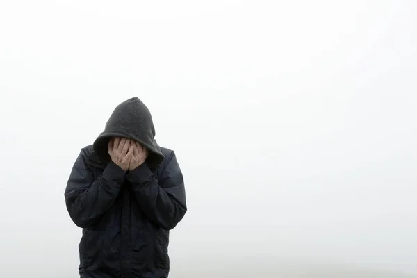 A hooded man holding his head in his hands. On a plain white background.