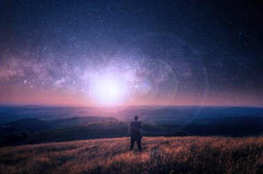 Yıldızdolu bir gökyüzü. Gökyüzünde parlak bir ışık ile manzara bakan bir tepeüzerinde bir yürüyüşçü.