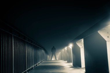 Sokak ışıkları yla siluetlenmiş bir patikada duran kapüşonlu bir figür