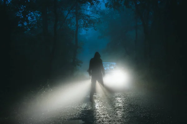 一个阴险的头戴头巾的人影站在一辆汽车前 在一个雾蒙蒙的夜晚 在一条恐怖的森林路上 — 图库照片