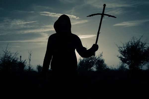 A spooky silhouette of a hooded man standing outside holding a wooden homemade cross at sunset. With a vintage grainy edit.
