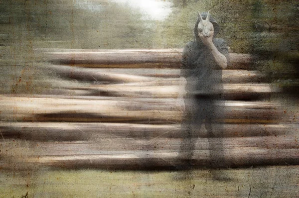 A scary, weird, hooded figure, holding a rabbit mask to his face. next to a pile of logs in a forest. With a blurred, grunge, vintage edit.