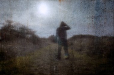 Bir ülke yolunda ki tarlada yalnız kapüşonlu bir figürün ürkütücü bir silueti. Güneşe bakıyor. Karanlık, ürkütücü bulanık soyut, grunge etkisi edit ile. 