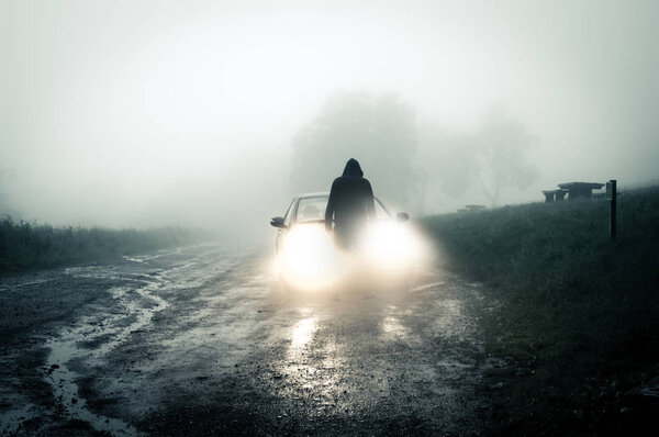 Одинокая, жуткая, в капюшоне фигура, стоящая перед машиной и смотрящая на пустую туманную просёлочную дорогу, силуэт которой был сделан ночью автомобильными фарами.
.