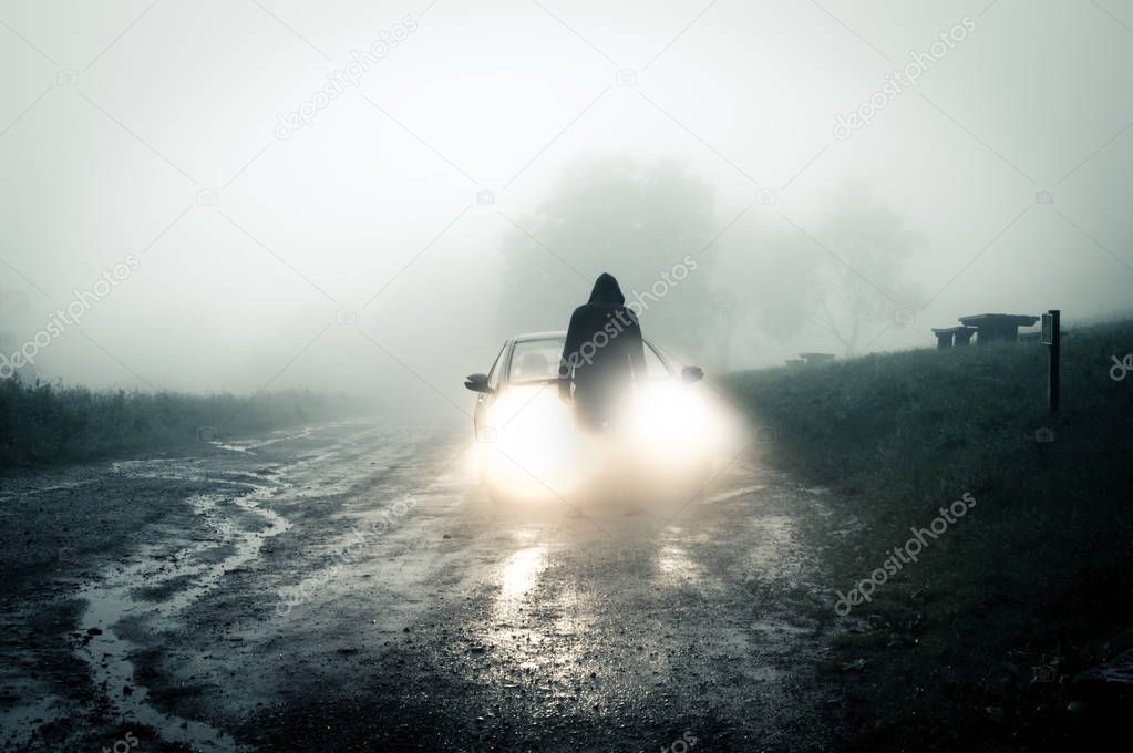 A lone, spooky, hooded figure standing in front of a car looking at an empty misty country road silhouetted at night by car headlights.