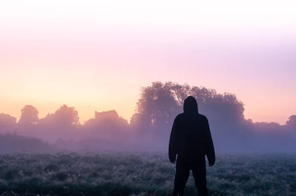 Eine geheimnisvolle einsame Kapuzenfigur, die an einem schönen frühen nebligen Morgen auf einem Feld steht und den Sonnenaufgang betrachtet. — Stockfoto