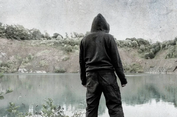 Mysterious hooded figure, standing and looking at a lake. With a grunge, vintsge edit.