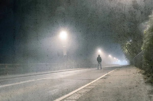 不気味な霧の夜に街路灯の下の道路の真ん中に立っている孤独なフード付きの人物 ヴィンテージで グランジ編集 — ストック写真