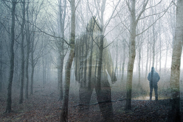 An artistic double exposure of a hooded figure standing in a forest on a spooky, foggy winters day. Looking at a man with a torch.