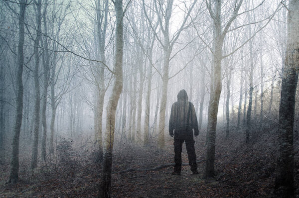 An artistic, double exposure of a hooded man standing in a spooky forest on a foggy winters day. With a grunge, blurred edit.