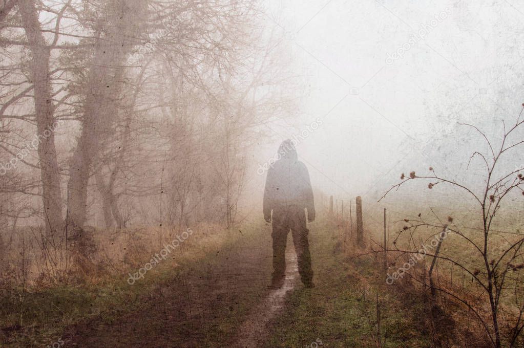 An artistic, double exposure of a hooded man standing on a spooky rural path on a foggy winters day. With a grunge, blurred edit.