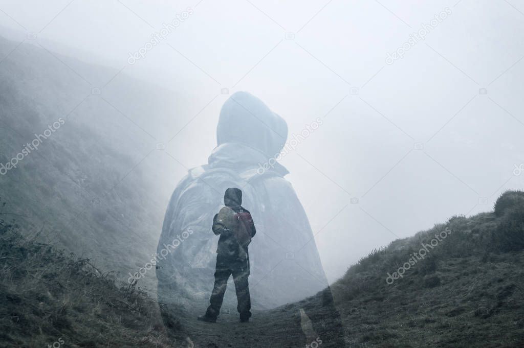 An artistic double exposure of a lone hooded hiker on a path in the countryside looking up on a cold spooky, foggy day.