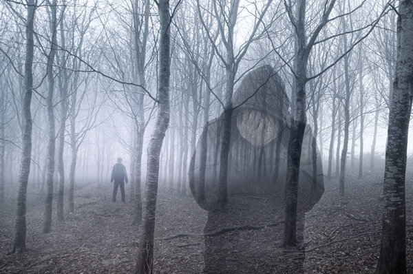 An artistic double exposure of a hooded figure holding his head in his hands, standing in a forest on a spooky, foggy winters day. With a figure standing in the background.