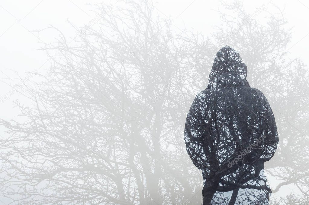 A double exposure, looking at the back of a hooded figure over layered by trees branches in winter.