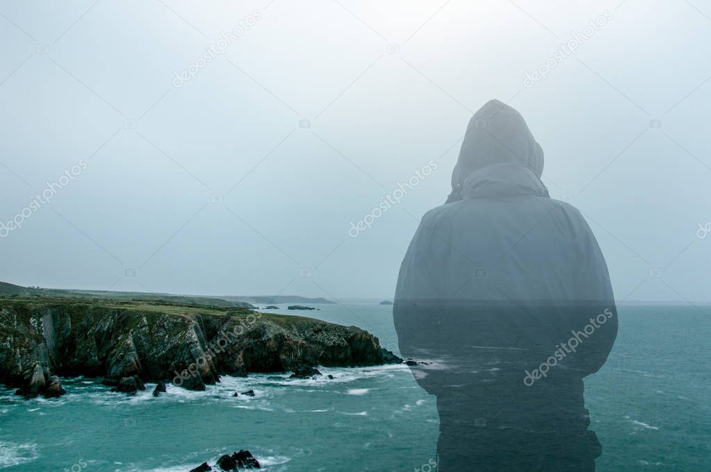 A double exposure of a hooded figure looking out at a moody rugged coast in winter.