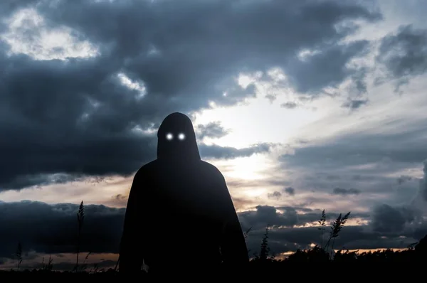 Um conceito de horror. Uma silhueta de uma figura encapuzada, de pé em um campo,, com olhos brilhantes assustadores olhando para a câmera. Voltar iluminado pelo sol poente . — Fotografia de Stock