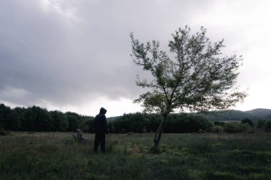 Ürkütücü bir kukuletalı figür, bir ağacın yanında duruyor. Fırtınalı bir akşamda kırsalda.