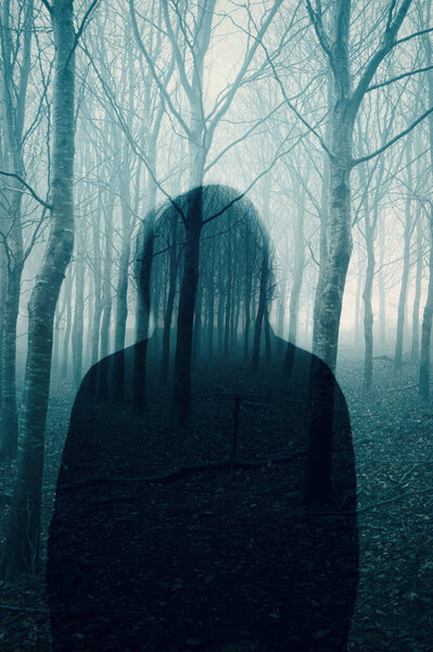 A double exposure of a Silhouette of a mysterious hooded figure without a face. Standing in a foggy winters forest