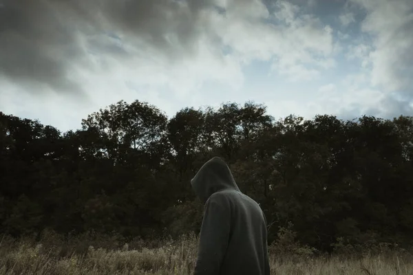 A moody, hooded figure, back to camera, standing in a field next to a forest.