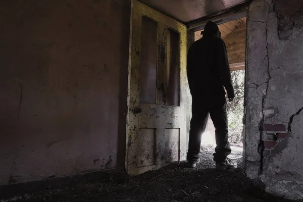 A hooded man, back to camera, standing in the doorway of a ruined, abandoned cottage.