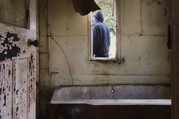 A dirty old bath tub in an abandoned cottsge in the countryside. With a hooded figure, back to camera.