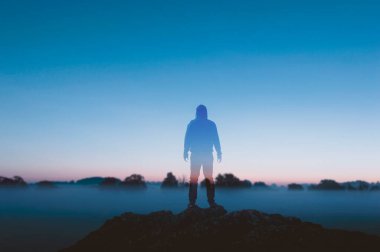 Sisli bir manzaranın odak dışı arka planına iki kez maruz kalmak. Tepede durup güneşin doğuşunu izleyen bir adamla..