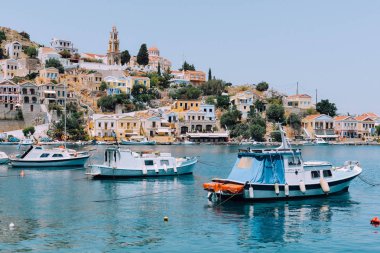 Symi Adası, Yunanistan - Haziran 2018 yaklaşık: güneşli bir günde, Yunanistan ve arka plan üzerinde renkli evleri sahil Symi Adası ve tepeler, berrak mavi suları tekneler float.