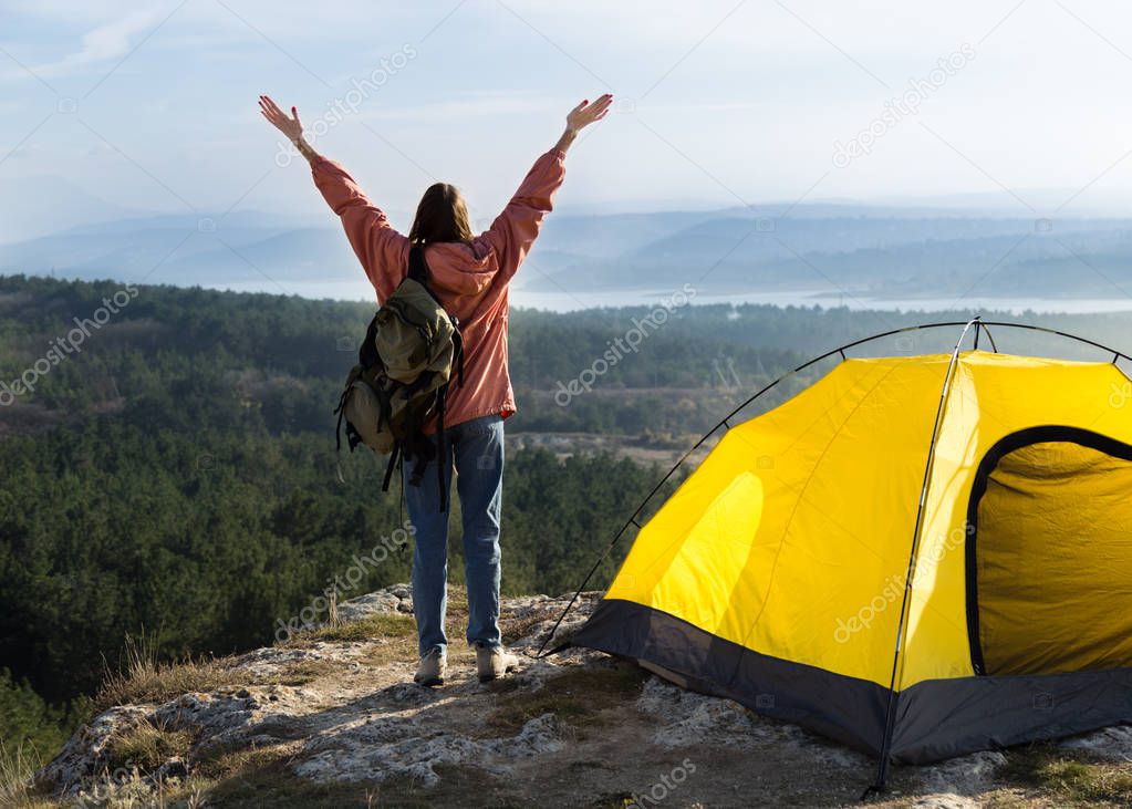 girl raises her hands to the sky against the backdrop of mountains, forests, lakes. Nearby is a tent. Real hiking photo.