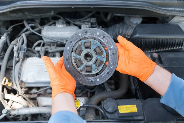 Auto mecánico con guantes de trabajo de protección sostiene el disco de embrague viejo por encima de un motor de coche —  Fotos de Stock