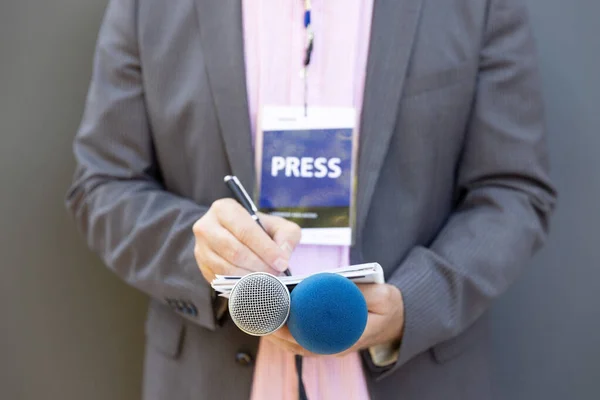 Male reporter at press conference or media event, writing notes, holding microphone