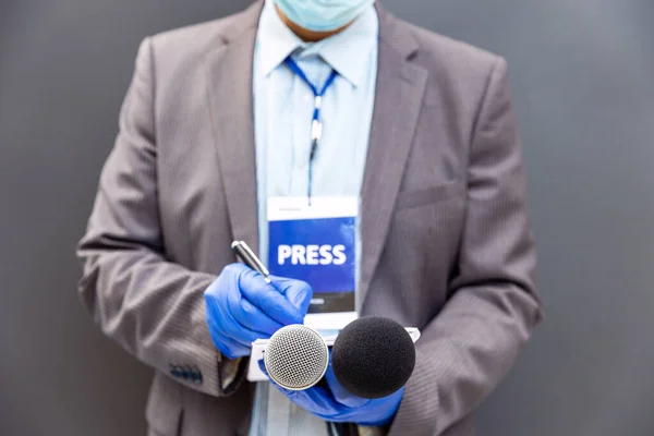 Journalist at news conference or media event wearing protective gloves and face mask against coronavirus COVID-19 disease holding microphone writing notes