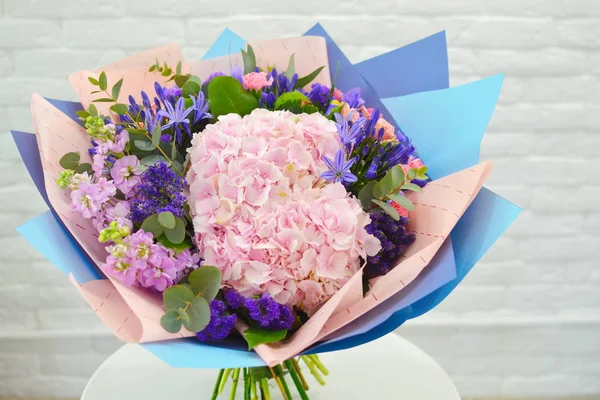 Beautiful lovely bouquet with hydrangea for flower shop. Beautiful bouquet of colorful flowers in packing on white table against the background of brick white wall. No people.  Close-up. Concept of flower shop.  Bouquet for catalog.