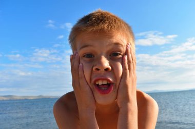 Şaşırmış yüzü olan sevimli komik çocuk. Genç ellerini yüzüne koydu. Temiz havada gölde ebeveynlerle yaz tatili. Yanlış anlama ve hayranlık samimi duygu.