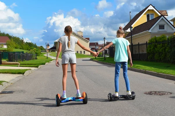 两个女朋友牵着一只手 女孩们骑在马路上的气垫板上 快乐快乐的暑假 孩子们牢固的友谊 快乐的孩子在夏天度假 — 图库照片