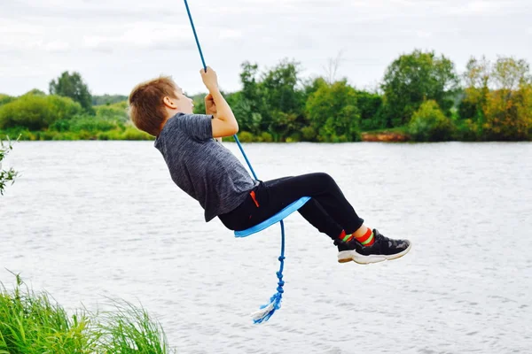 Cheerful boy swinging on a rope swing above the water. — Stock Photo, Image
