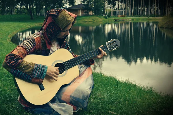 Una donna hipster suona la chitarra sull'erba vicino al lago. Retro — Foto Stock