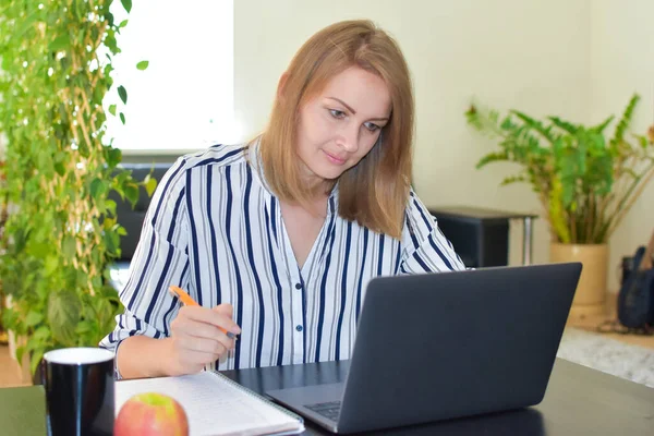 Жінка з ноутбуком онлайн урок пише нотатки за столом у кімнаті — стокове фото